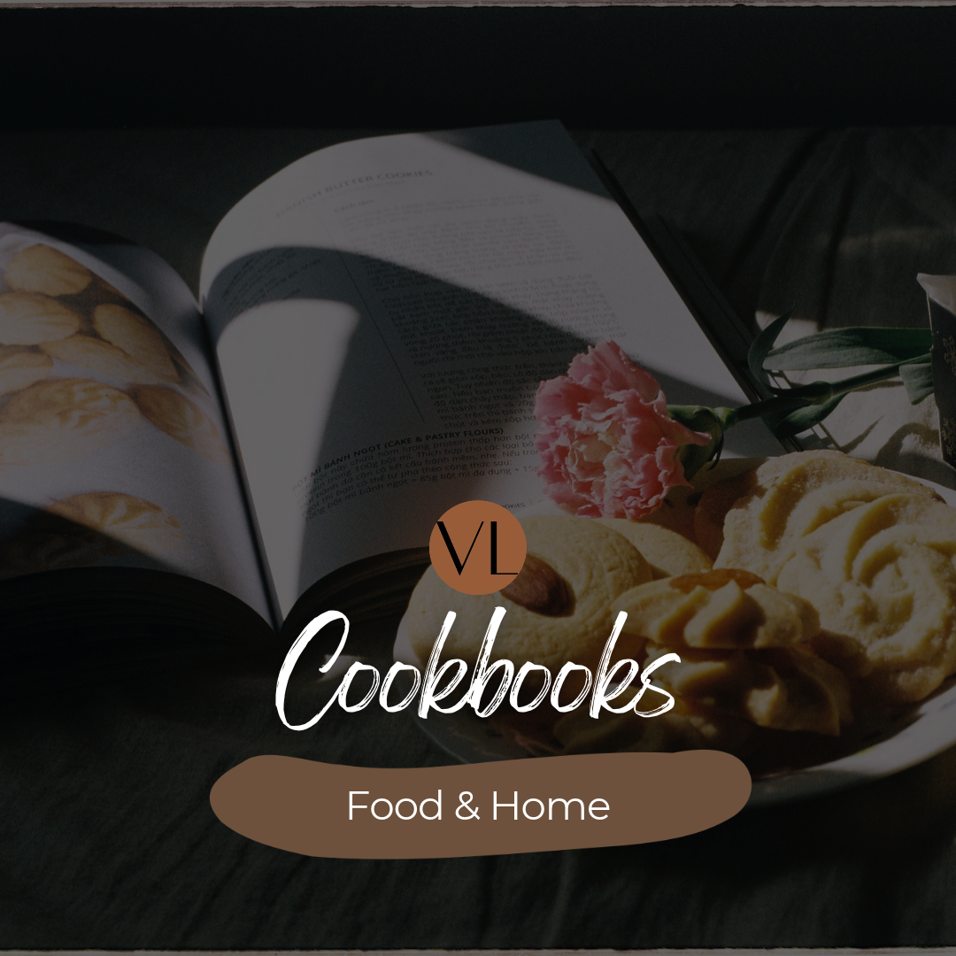 A dark-themed promotional graphic for a cookbook titled 'VL Cookbooks Food & Home', featuring an open book with recipe pages and a plate of cookies with a pink flower on a dark wooden surface.